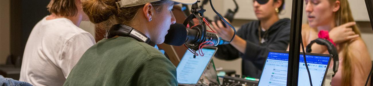 Four students at a podcasting station with the closest student talking into a microphone