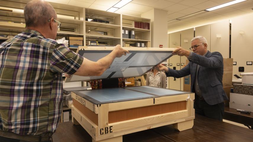 Two people lift the lid of a wood crate to unpack the diploma.