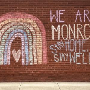 Brick wall with a chalk rainbow and the words "We are Monroe. Stay Home. Stay well"
