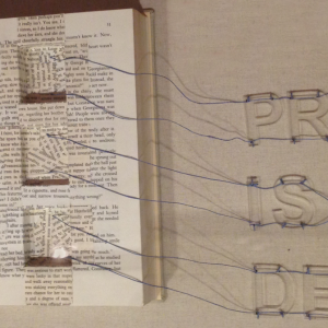 Open book with three square sections cut out. Strings from the cut-outs spell the words Print is Dead