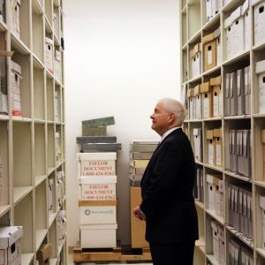 Robert Gates frames by two sets of shelves containing his papers in the Special Collections archive
